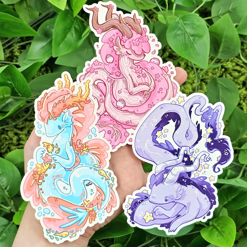 May include: Three colorful stickers featuring whimsical, cartoon-style sea creatures. The first sticker is pink and features a creature with antlers and a floral design. The second sticker is blue and features a creature with a tail and seaweed. The third sticker is purple and features a creature with a star-shaped tail and a starry design.