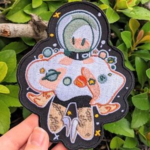 May include: Embroidered patch featuring a cartoon astronaut in a white spacesuit with a green helmet, surrounded by stars and planets. The astronaut is wearing a black and white outfit with brown boots. The patch is on a black background.