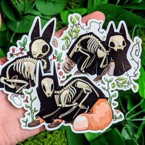 May include: Three black and white skeleton animal stickers with a spooky, gothic aesthetic. Each sticker features a different animal wearing a black cloak with a hood. The animals are a fox, a cat, and a rabbit.