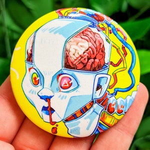 May include: A yellow button with a cartoon image of a robot with a red and white brain exposed. The robot has a blue and yellow wire coming out of its head and a red nose. The robot's eyes are red with a white triangle in the center.