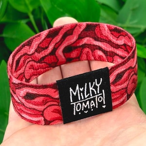 May include: A red and black patterned elastic hair tie with the text "MILKY TOMATO!" on a black tag. The hair tie is a vibrant red with a swirling, abstract design.