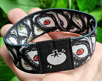 May include: Black and white wristband with a repeating pattern of eyes and a black buckle with a white tomato graphic. The buckle has the text "Shroom Queens" on it.