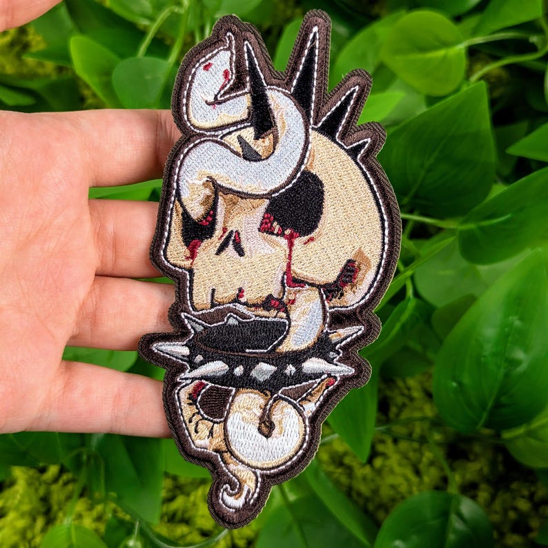 May include: A white snake wrapped around a brown skull with a black and silver studded collar. The skull has red paint dripping from the eye sockets.