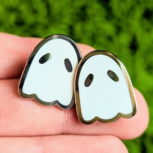 May include: Two white ghost-shaped enamel pins with gold outlines. The pins are on a hand.