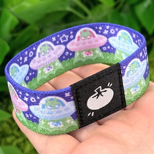May include: A blue, green, and purple patterned bracelet featuring cartoon UFOs and stars. The bracelet has a black tag with a white graphic of a tomato. The bracelet is approximately 1 inch wide.