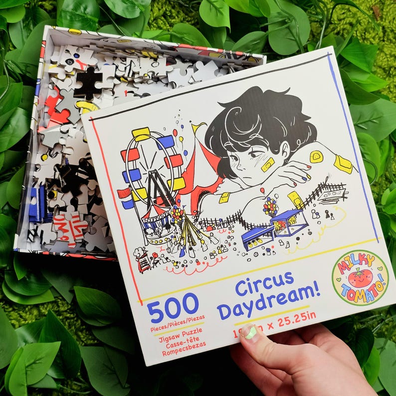 May include: A 500-piece jigsaw puzzle featuring a colourful illustration of a circus scene with a Ferris wheel, a person with a sad expression, and the text "Circus Daydream!" and "MILKY TOMATO".
