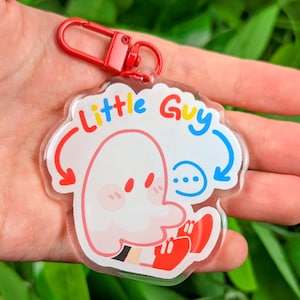 May include: A clear acrylic keychain with a red clasp. The keychain features a cartoon ghost character with the text "Little Guy" in colorful letters. The ghost has red shoes and blush marks on its cheeks.