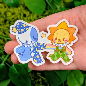 May include: Two die-cut stickers featuring cartoon characters. One is a blue clown with a star-shaped hat and the other is a smiling sun with a rainbow arm. The stickers are held in a hand against a green background.
