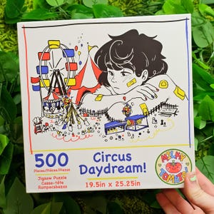 May include: A 500-piece jigsaw puzzle featuring a colourful illustration of a circus scene with a Ferris wheel, a tent, and a person daydreaming. The puzzle is titled "Circus Daydream!" and is made by Milky Tomato. The puzzle measures 49.5 cm by 64.1 cm.