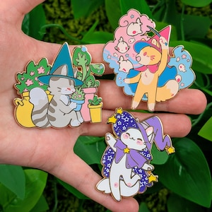 May include: Three enamel pins featuring cartoon cats in wizard attire. One cat is tending to plants, another is casting a spell, and the third is in a purple cape and hat. Each pin has gold-colored metal accents.