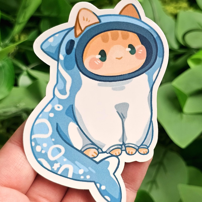 Puede incluir: Una pegatina de un gato de dibujos animados con un disfraz de tibur&oacute;n azul. El gato tiene pelaje naranja y ojos verdes. El disfraz de tibur&oacute;n tiene una barriga blanca y escamas azules.