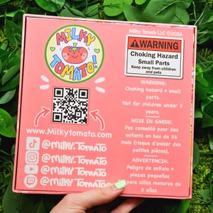 May include: Pink box with a cartoon tomato logo and the text "Milky Tomato". The box has a QR code, social media handles, and a warning label about choking hazards for children under 3 years old.