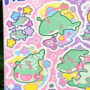 Puede incluir: Una hoja de pegatinas con ilustraciones caprichosas con tem&aacute;tica espacial. Las pegatinas muestran criaturas verdes con forma de tibur&oacute;n, cohetes, planetas y estrellas en colores pastel sobre un fondo rosa. Los dise&ntilde;os tienen un estilo juguet&oacute;n y de dibujos animados.