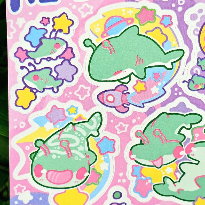 Puede incluir: Una hoja de pegatinas con ilustraciones caprichosas con tem&aacute;tica espacial. Las pegatinas muestran criaturas verdes con forma de tibur&oacute;n, cohetes, planetas y estrellas en colores pastel sobre un fondo rosa. Los dise&ntilde;os tienen un estilo juguet&oacute;n y de dibujos animados.