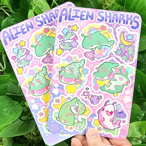 Puede incluir: Dos hojas de pegatinas con el texto "ALIEN SHARKS" en morado. Cada hoja presenta varias ilustraciones de tiburones alienígenas de dibujos animados en verde y rosa, junto con estrellas, planetas y cohetes sobre un fondo rosa.