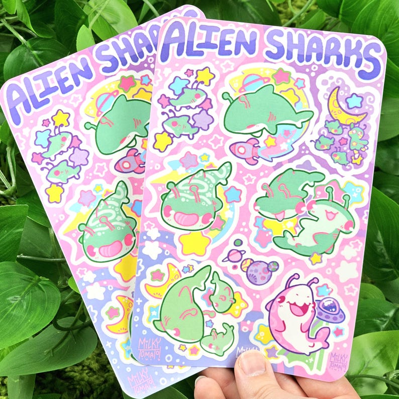 Puede incluir: Dos hojas de pegatinas con el texto "ALIEN SHARKS" en morado. Cada hoja presenta varias ilustraciones de tiburones alien&iacute;genas de dibujos animados en verde y rosa, junto con estrellas, planetas y cohetes sobre un fondo rosa.