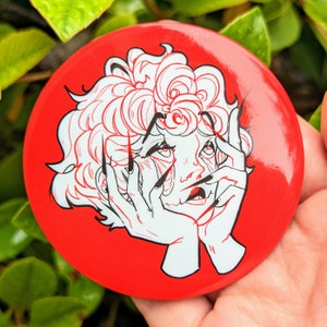 May include: A red round button with a white line drawing of a person with curly hair, their hands covering their face. The person's eyes are wide open and their mouth is slightly open. The hands have long, sharp nails.
