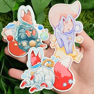 May include: Four colorful stickers featuring cartoon foxes in astronaut suits. The foxes are depicted in various poses, surrounded by planets, stars, and space-themed elements. The stickers have a whimsical and playful design.