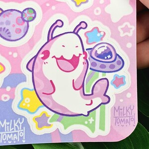 Puede incluir: Una pegatina con un personaje alien&iacute;gena de dibujos animados en rosa y blanco, sonriendo. El fondo es rosa con estrellas, planetas y un OVNI. La pegatina tiene el texto "Milky Tomato" impreso.
