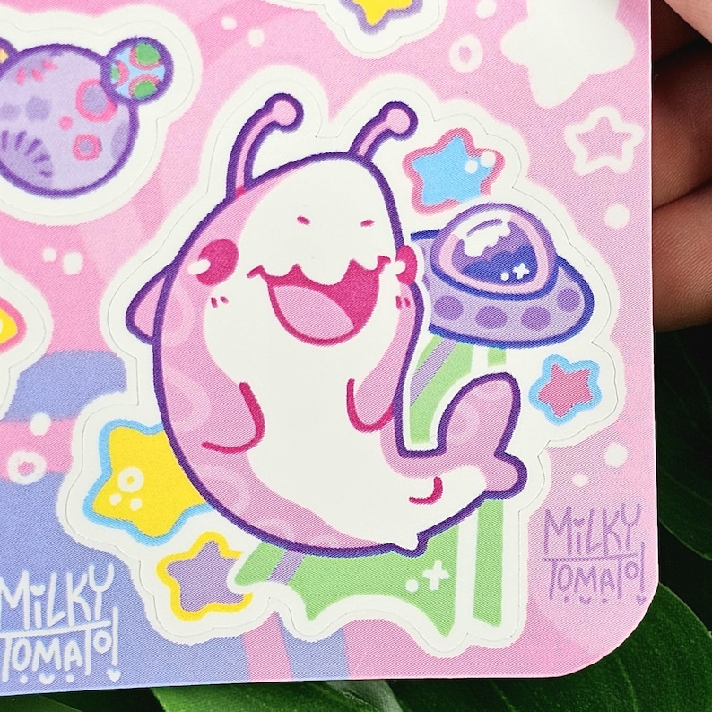 Puede incluir: Una pegatina con un personaje alien&iacute;gena de dibujos animados en rosa y blanco, sonriendo. El fondo es rosa con estrellas, planetas y un OVNI. La pegatina tiene el texto "Milky Tomato" impreso.