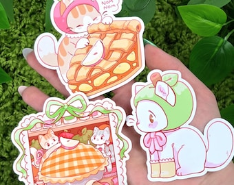 Könnte beinhalten: Drei farbenfrohe Aufkleber mit Cartoon-Katzen. Eine Katze trägt einen rosa Hut und isst Apfelkuchen, mit einer Sprechblase mit der Aufschrift "NUM NOM NOM". Ein weiterer Aufkleber zeigt Katzen mit einem Kuchen. Die dritte Katze trägt einen grünen Hut.
