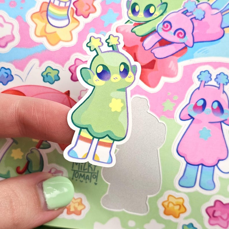 May include: A green alien sticker with a star on its chest and rainbow socks. The sticker is on a pink and green background with other stickers of aliens and stars.