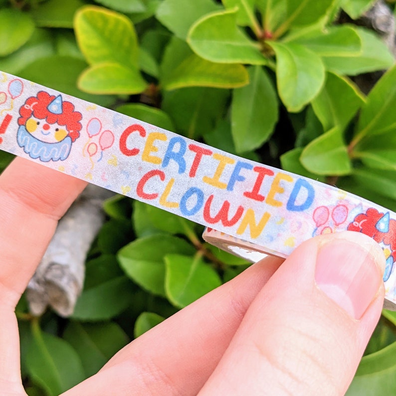May include: A white washi tape with the text "CERTIFIED CLOWN" in rainbow colours. The tape also features a cartoon clown with a red hat and a red nose. There are also small balloons and confetti on the tape.