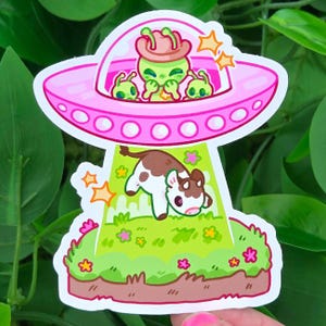 May include: A sticker featuring a pink UFO with green aliens wearing a cowboy hat, beaming up a brown and white cow. The scene is set on a green grassy field with flowers and stars.