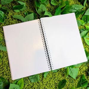 May include: An open, blank spiral-bound notebook with white pages. The notebook is positioned on a green, textured surface, surrounded by green leaves. The spiral binding is black and white.