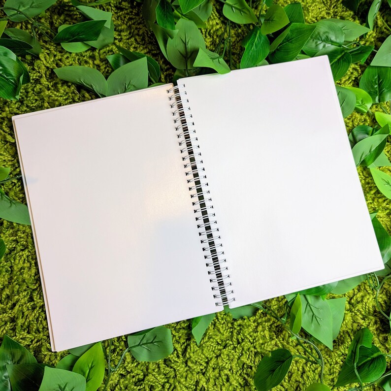 May include: An open, blank spiral-bound notebook with white pages. The notebook is positioned on a green, textured surface, surrounded by green leaves. The spiral binding is black and white.