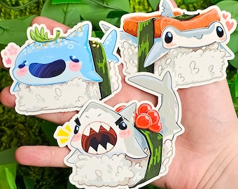 May include: Three colorful shark sushi stickers. Each sticker features a different shark species with a seaweed and rice design. The sharks are a blue and white hammerhead, a gray reef shark, and a blue and white whale shark.