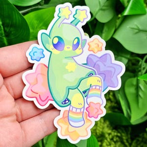 May include: A green alien-like creature with large eyes and a star on its chest sits on a bed of colorful stars. The creature is wearing rainbow striped socks and has a yellow and green body.