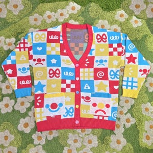 May include: A vibrant, patterned cardigan. The V-neck sweater has long sleeves and a patchwork design with red, yellow, blue, and white squares, decorated with cartoon graphics. It has a red trim and buttons. The measurements are in cm.