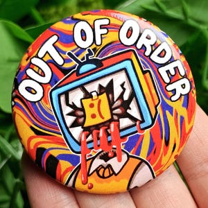 May include: A colorful button with a swirling pattern in red, yellow, blue, and white. The text "OUT OF ORDER" is written in white around the edge of the button. A cartoon television with a sad face is in the center of the button.