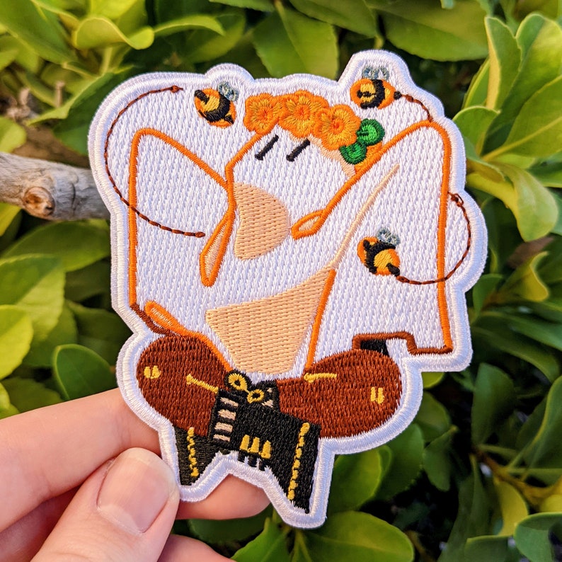 May include: White embroidered patch with a cartoon character wearing a brown and orange outfit and a crown of orange flowers. The character is surrounded by three yellow and black bees.