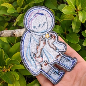 May include: A white and blue embroidered patch of a cartoon astronaut holding a star. The astronaut is wearing a helmet and a spacesuit. The patch is perfect for adding a whimsical touch to any project.