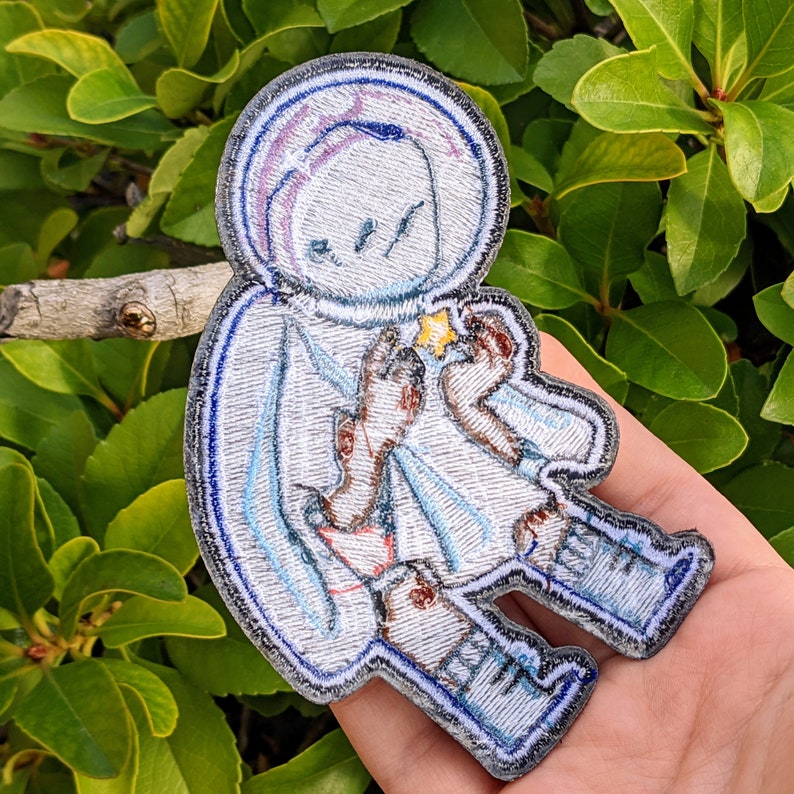 May include: A white and blue embroidered patch of a cartoon astronaut holding a star. The astronaut is wearing a helmet and a spacesuit. The patch is perfect for adding a whimsical touch to any project.