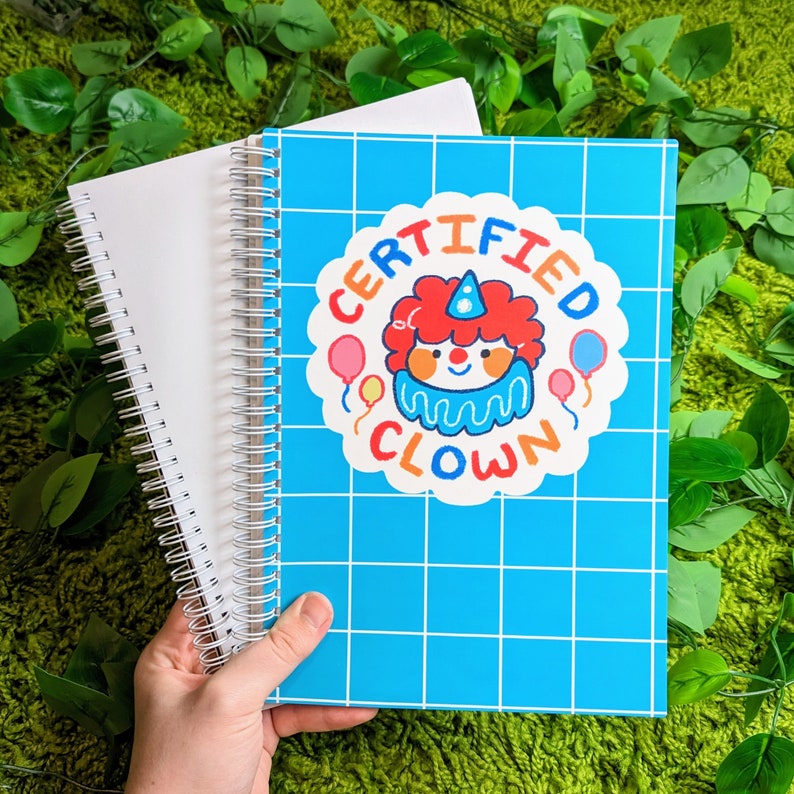 May include: Blue spiral bound notebook with a white grid pattern and a colorful sticker that says "Certified Clown". The sticker features a cartoon clown with a red hat and a blue and white striped shirt.