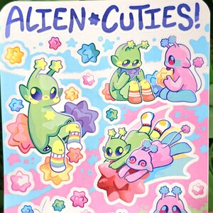 May include: A sheet of stickers featuring cute alien characters with colorful star accents. The stickers have a pink, blue, and white background with the text "ALIEN*CUTIES!" at the top.