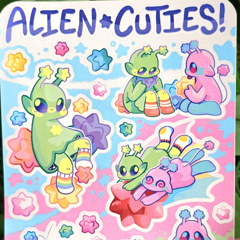 May include: A sheet of stickers featuring cute alien characters with colorful star accents. The stickers have a pink, blue, and white background with the text "ALIEN*CUTIES!" at the top.