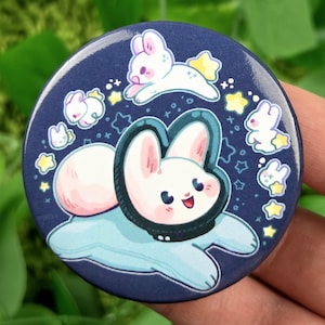 May include: A circular pin-back button featuring a dark blue background with a central illustration of a white rabbit wearing a black space helmet. The design includes several smaller white rabbits and yellow stars scattered around the centre.