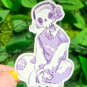 Puede incluir: Una pegatina morada y blanca de un esqueleto de dibujos animados que lleva un su&eacute;ter morado y auriculares con una flor en la parte superior. El esqueleto est&aacute; sentado con las piernas cruzadas.