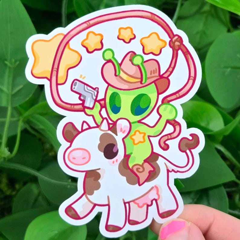 May include: A sticker featuring a green alien wearing a cowboy hat and holding a gun, riding a cow. The cow is white with brown spots. A lasso and yellow stars surround the alien. The sticker has a pink outline.