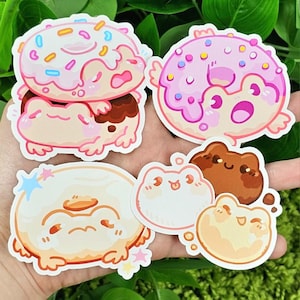 May include: A selection of vibrant stickers showcasing cartoon frogs with donut designs. The stickers feature pink, brown, and white colours, with sprinkles and star accents. The frog stickers are in various poses and sizes.