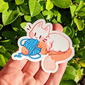 K&ouml;nnte beinhalten: Eine wei&szlig;e Katze mit rosa Akzenten ist mit einem blauen Wollkn&auml;uel zusammengerollt. Die Katze hat einen niedlichen, cartoonartigen Stil und h&auml;lt das Garn mit ihren Pfoten.