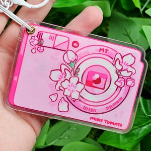May include: Pink camera-shaped acrylic keychain with a white outline and a pink and white cherry blossom design. The keychain has the text "MT Milky Tomato" on the bottom.