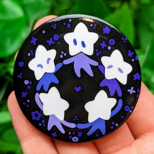 May include: A black button with a white background featuring five white stars with purple outlines holding hands in a circle. The stars have small black eyes and mouths. The background is filled with small white and purple dots and stars.