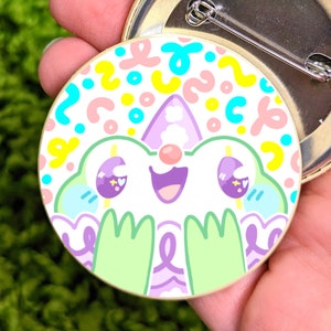 May include: A colourful pin featuring a cartoon character with a happy expression. The character has large eyes, a pink nose, and green arms. The background is a white circle with a colourful abstract pattern.