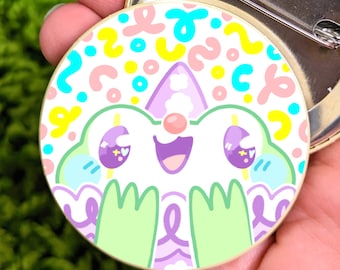 May include: A colorful pin featuring a cartoon character with a happy expression. The character has large eyes, a pink nose, and green arms. The background is a white circle with a colorful abstract pattern.