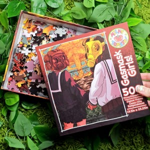 May include: A 500-piece jigsaw puzzle box featuring two girls wearing gas masks. The girls are standing in front of a large explosion. The box is red and white with the text "Milky Tomato Gasmask Girls!" and "19.5in x 25.25in".